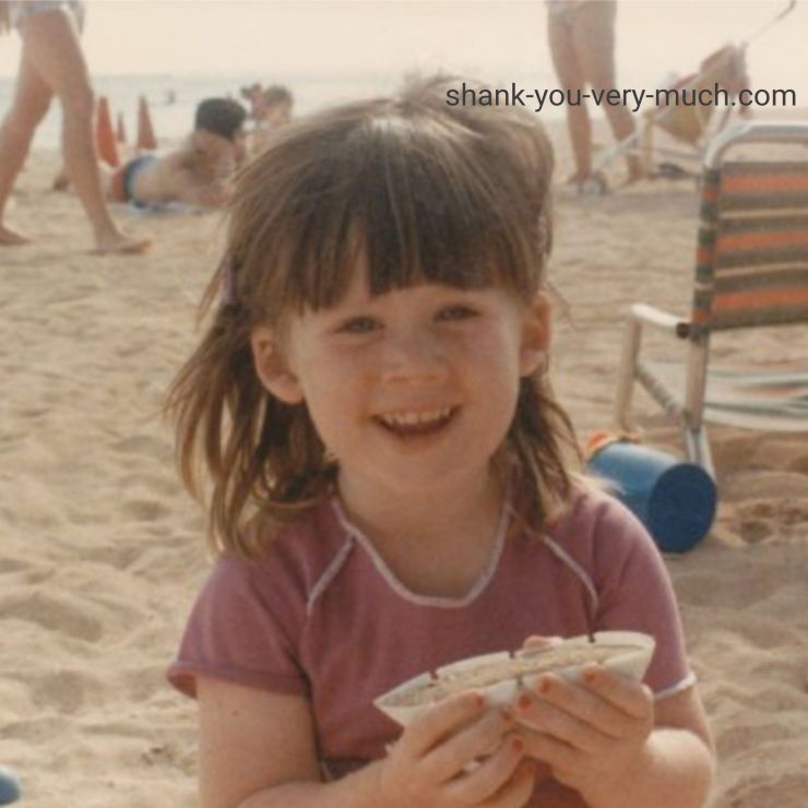 A photo of young Heather sitting on a sandy beach smiling.